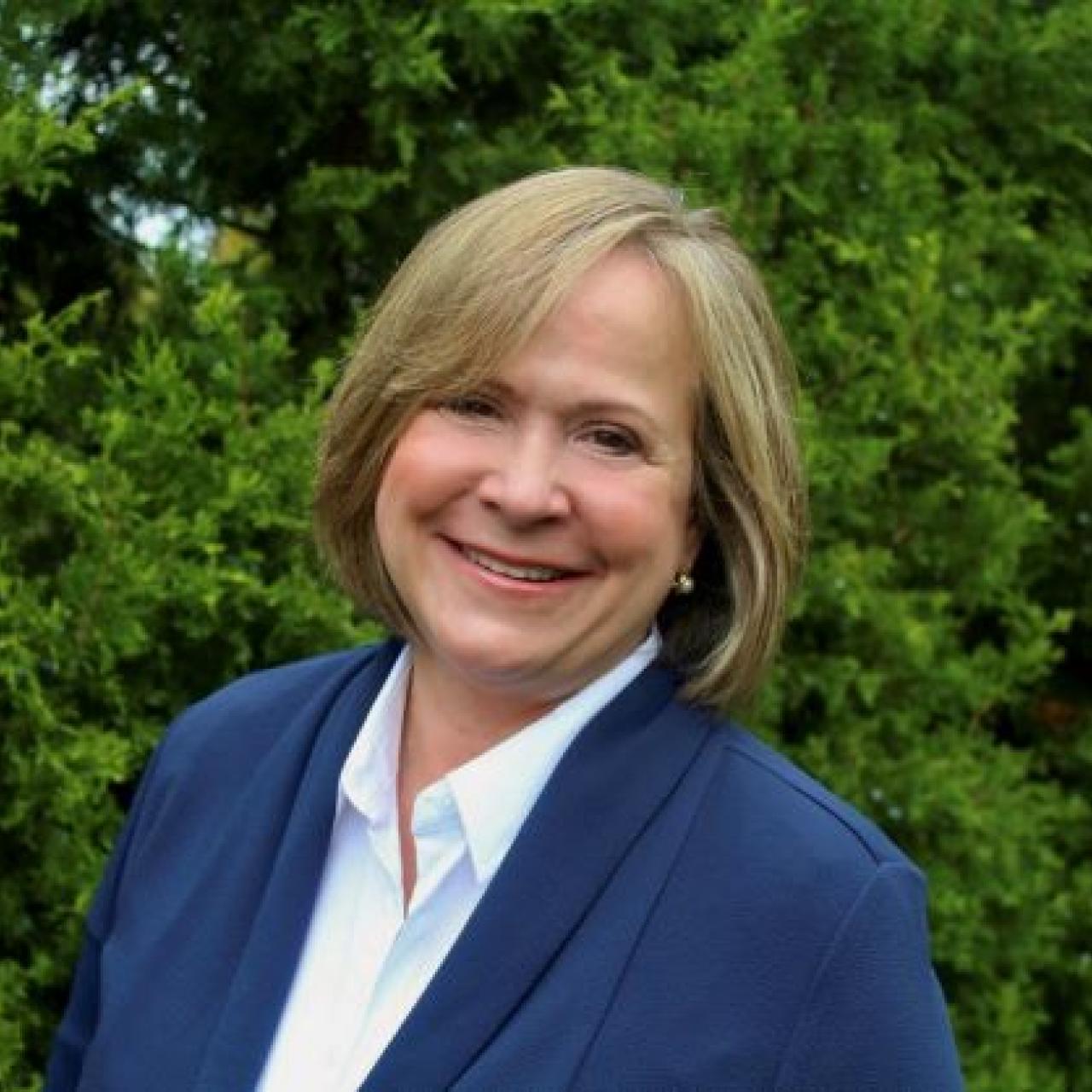 Arlene headshot in front of trees wearing a blue blazer and white collared shirt.