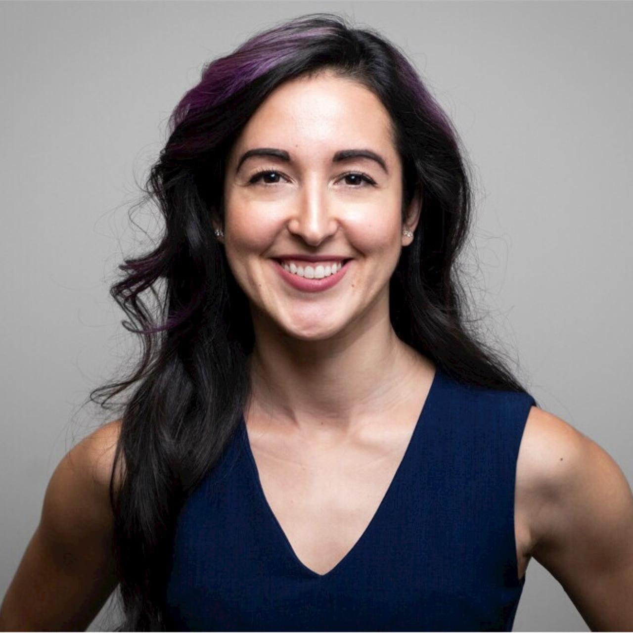 Image of Amelia smiling into camera with blue v-neck blouse against gray background.