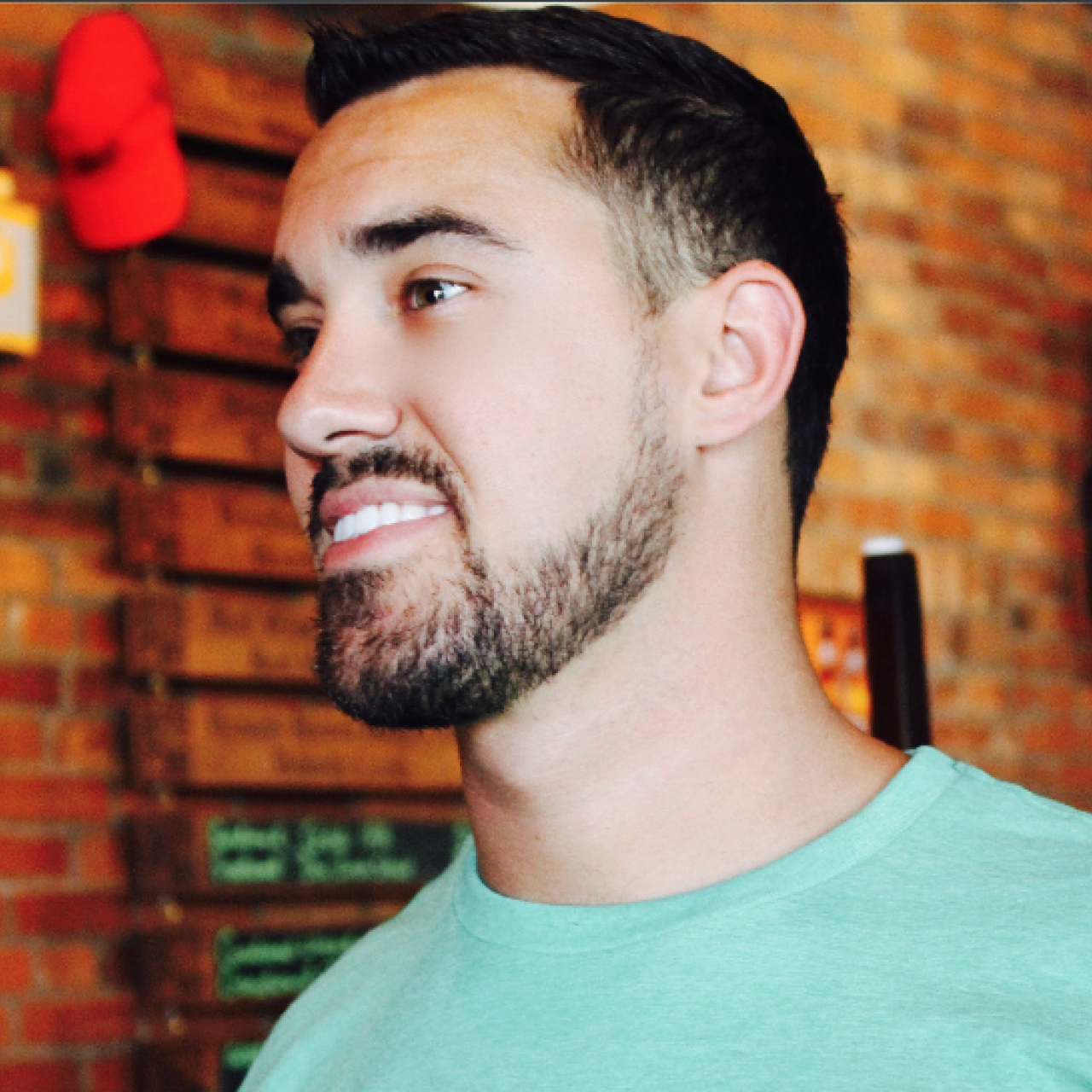 Image of Trevor in a green shirt looking off to the side, standing in front of brick wall.