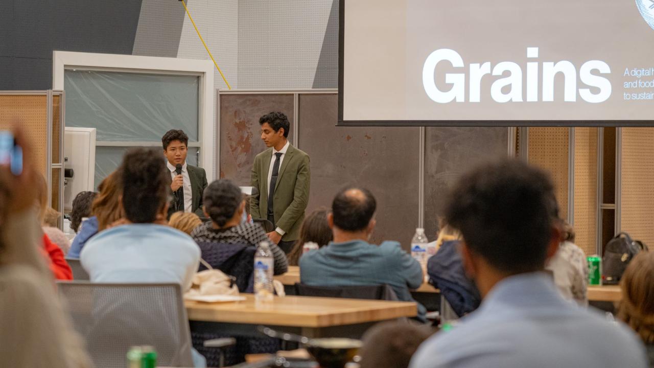 Two members of grains team presenting at front of room with screen and wearing suits